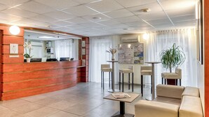 Zona de recepción de hotel con un mostrador de madera, un área de espera con sofá y sillones, y una pequeña mesa con un periódico.