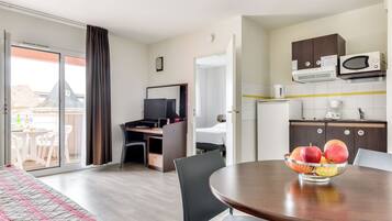 Habitación de hotel con cama, escritorio con computadora, mesa de comedor con fruta, área de cocina con armarios y electrodomésticos, y puerta corrediza de vidrio a un balcón.
