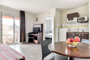 A hotel room with a bed, a desk with a computer, a dining table with fruit, a kitchen area with cabinets and appliances, and a sliding glass door to a balcony. - Zenitude Hôtel Résidences Beziers Centre (Béziers)