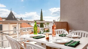 Mesa en balcón con platos, vasos y botellas, con vista a un paisaje urbano con edificios y un árbol.