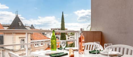 A balcony table set with plates, glasses, and bottles, overlooking a cityscape with buildings and a tree.