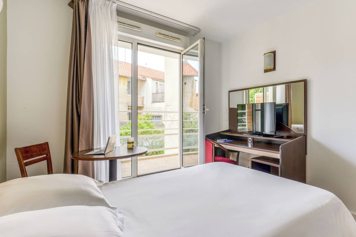 A hotel room with a bed, a desk with a computer, a chair, a TV, and a sliding glass door to an outdoor balcony.