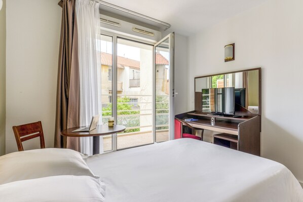 A hotel room with a bed, a desk with a computer, a chair, a TV, and a sliding glass door to an outdoor balcony.