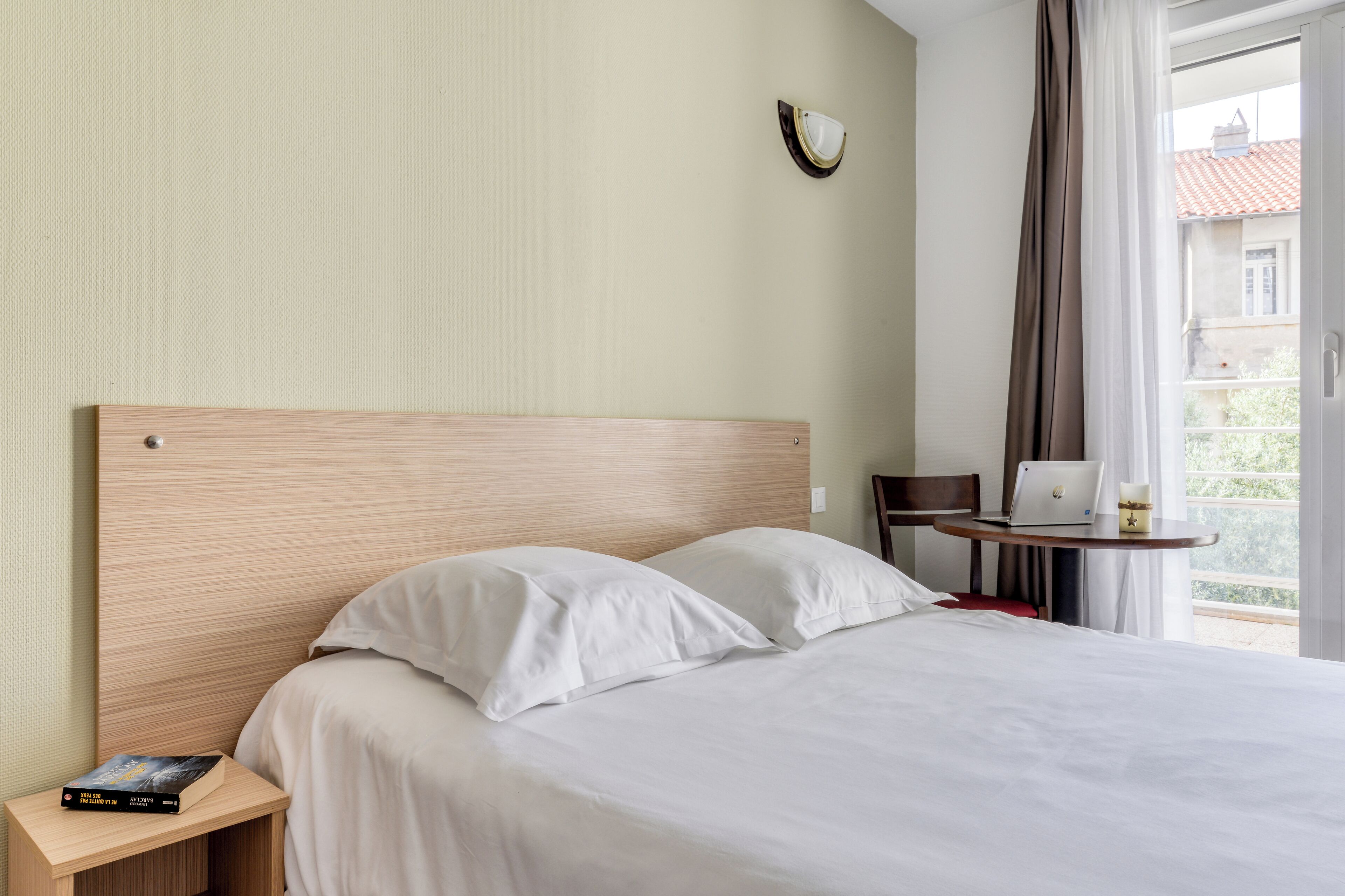 Hotellihuone, jossa on sänky, yöpöytä, jossa on kirja, kannettava tietokone pienellä pöydällä ja näkymä ulos.
