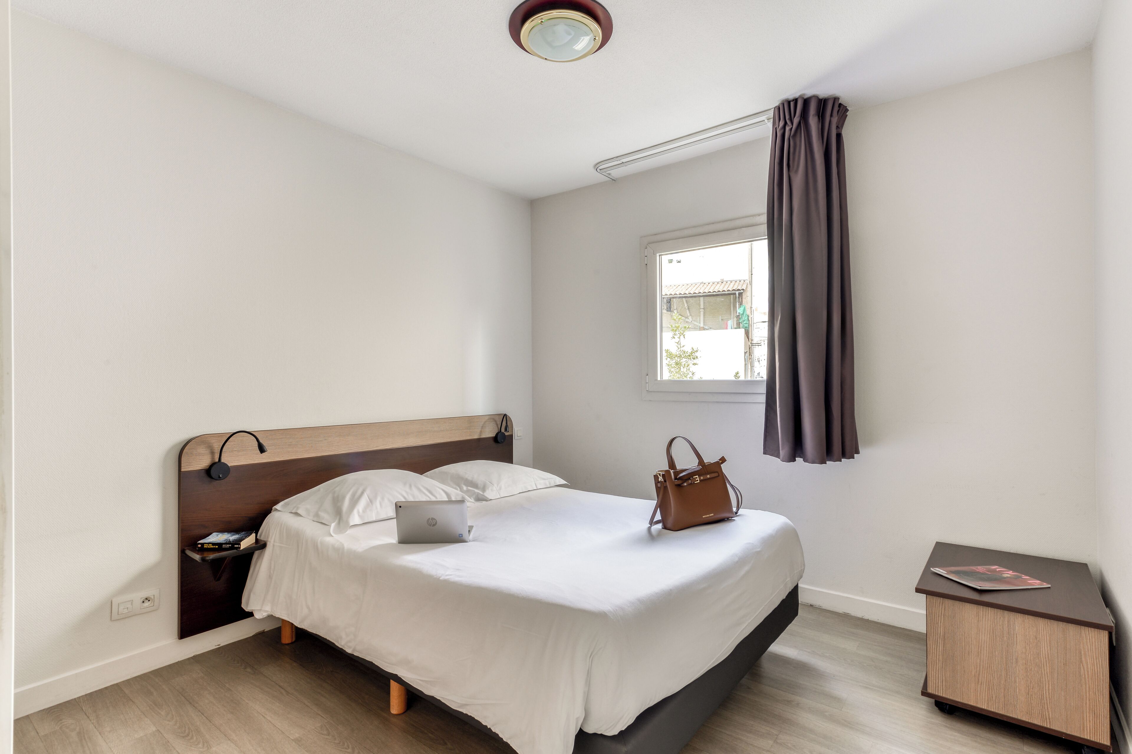 A neatly arranged bedroom with a bed, a nightstand, a laptop, a handbag, and a small wooden cabinet.