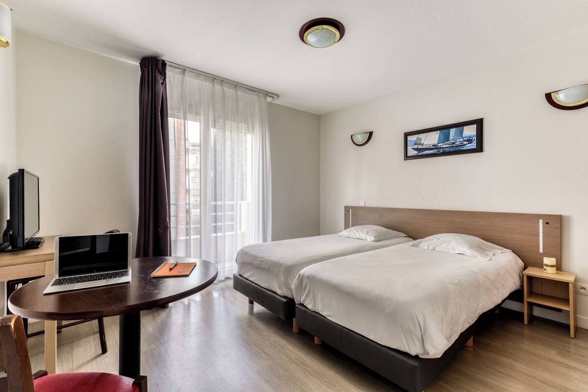 A hotel room with a bed, a desk with a laptop, a chair, a bedside table with a lamp, and a window with curtains.