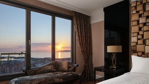 Habitación de hotel con un ventanal grande que ofrece vista a la ciudad al atardecer, un sofá, una mesita de noche con lámpara y una cama con colcha a rayas.