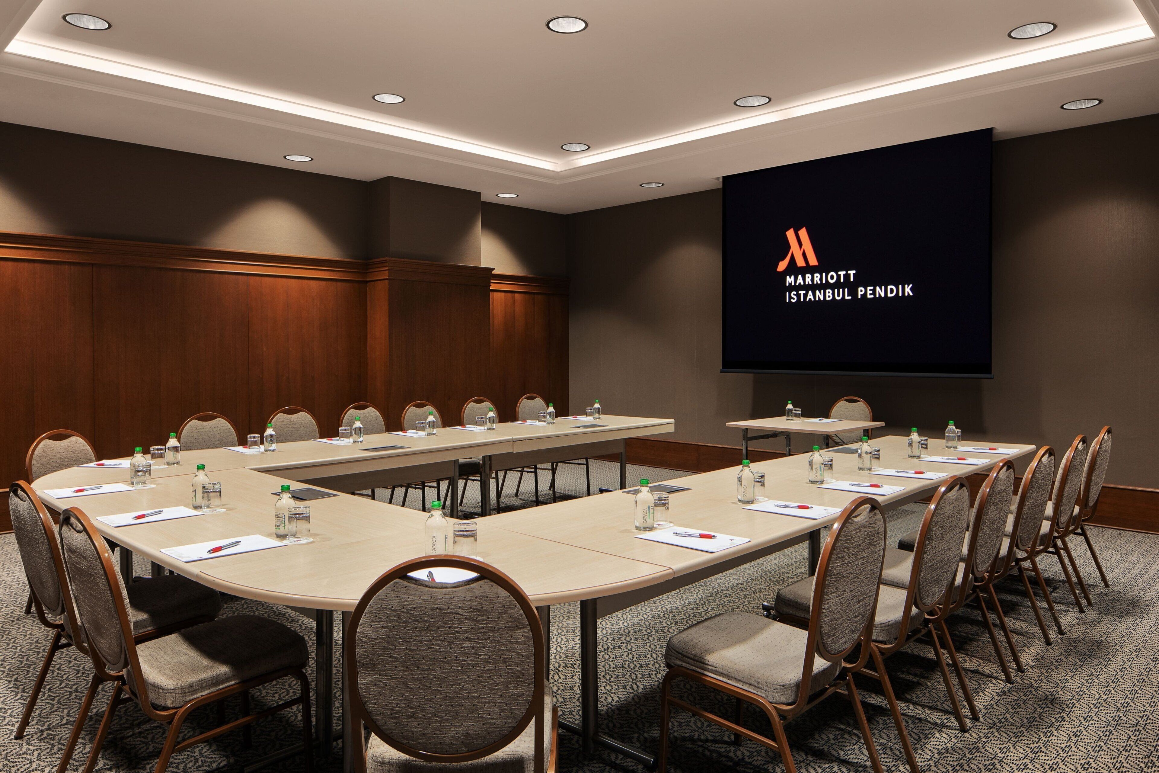 Ein Konferenzraum mit einem langen Tisch, Stühlen und einem Bildschirm, der das Logo des Marriott Istanbul Pendik zeigt.