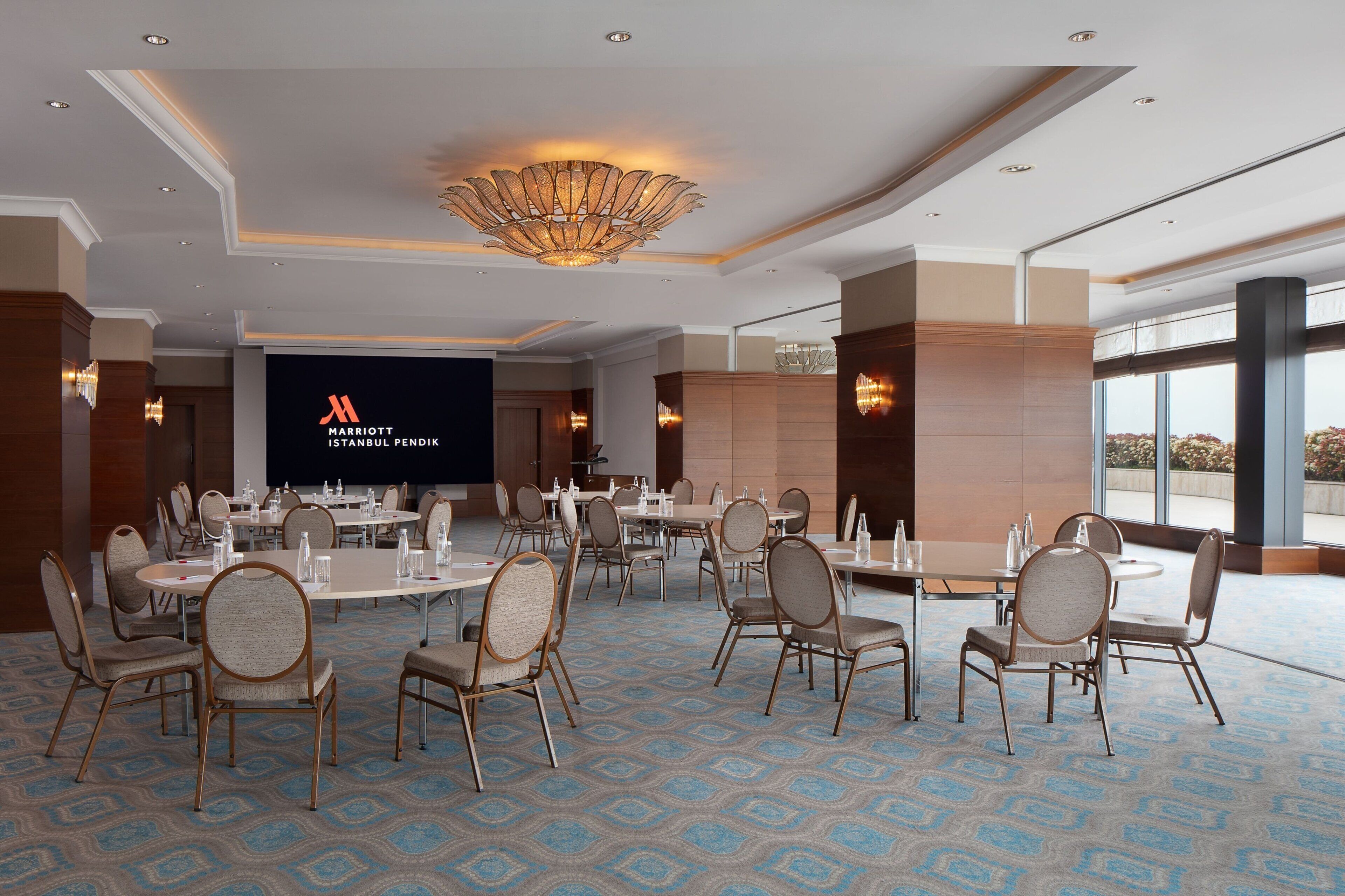 A conference room with a large screen displaying the Marriott Istanbul logo, arranged tables and chairs, and a patterned carpet.
