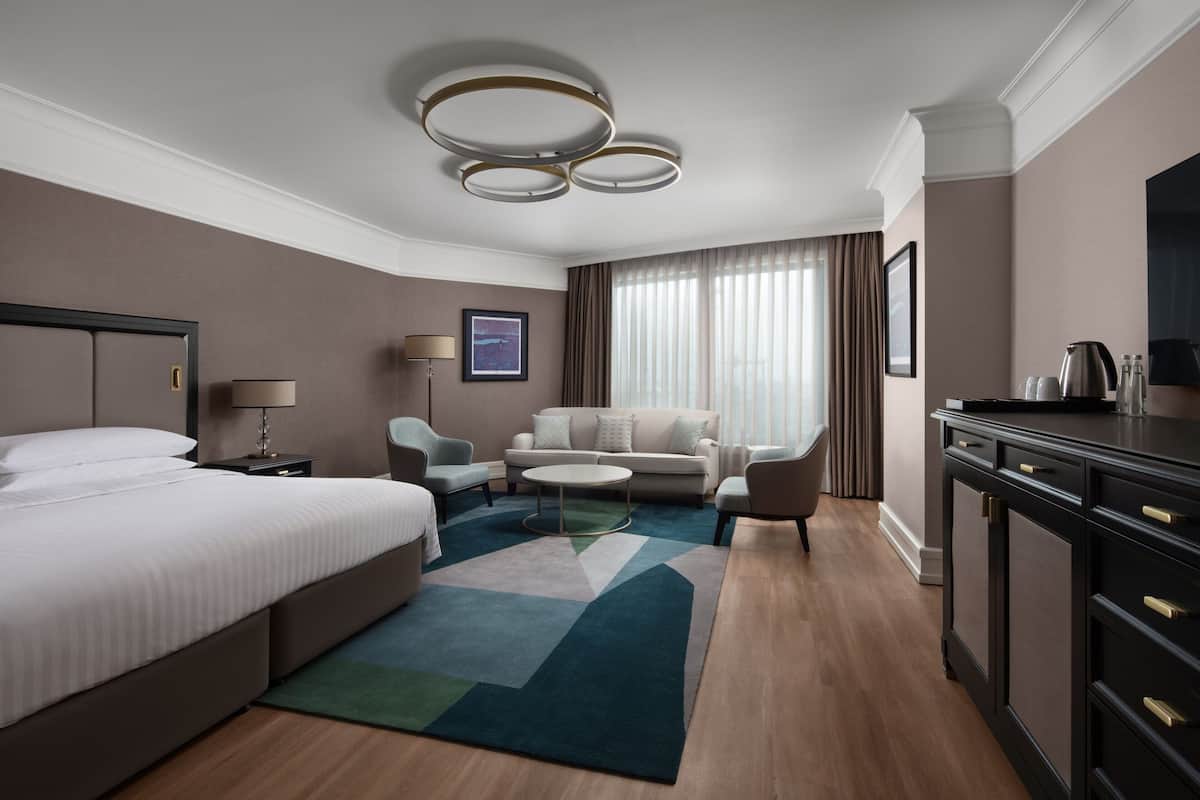 A modern hotel room with a large bed, a sofa, a coffee table, and a desk with drawers.