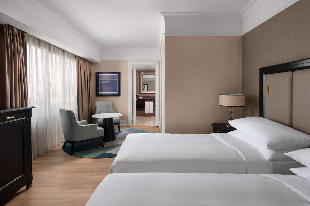 A hotel room with two beds, a desk, a chair, a lamp, and a view of the hallway.