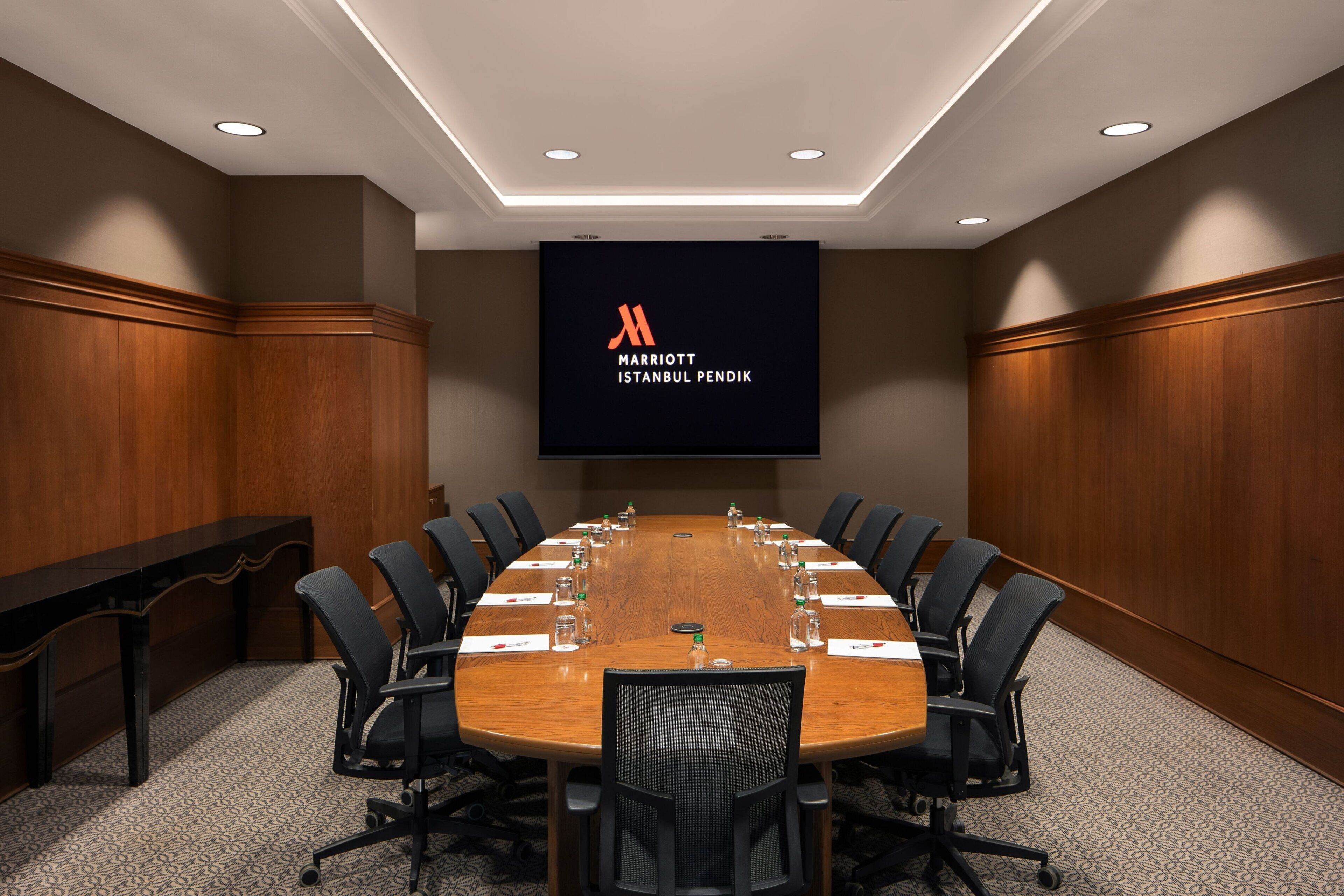 Ett konferensrum med ett långt bord i trä, svarta stolar och en stor skärm som visar Marriott Istanbul Pendiks logotyp.