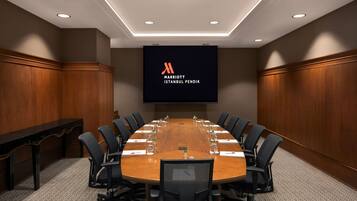Ett konferensrum med ett långt bord i trä, svarta stolar och en stor skärm som visar Marriott Istanbul Pendiks logotyp.