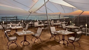 Un área de comedor en la azotea con mesas y sillas, grandes sombrillas blancas y vistas a la ciudad y al agua al atardecer.