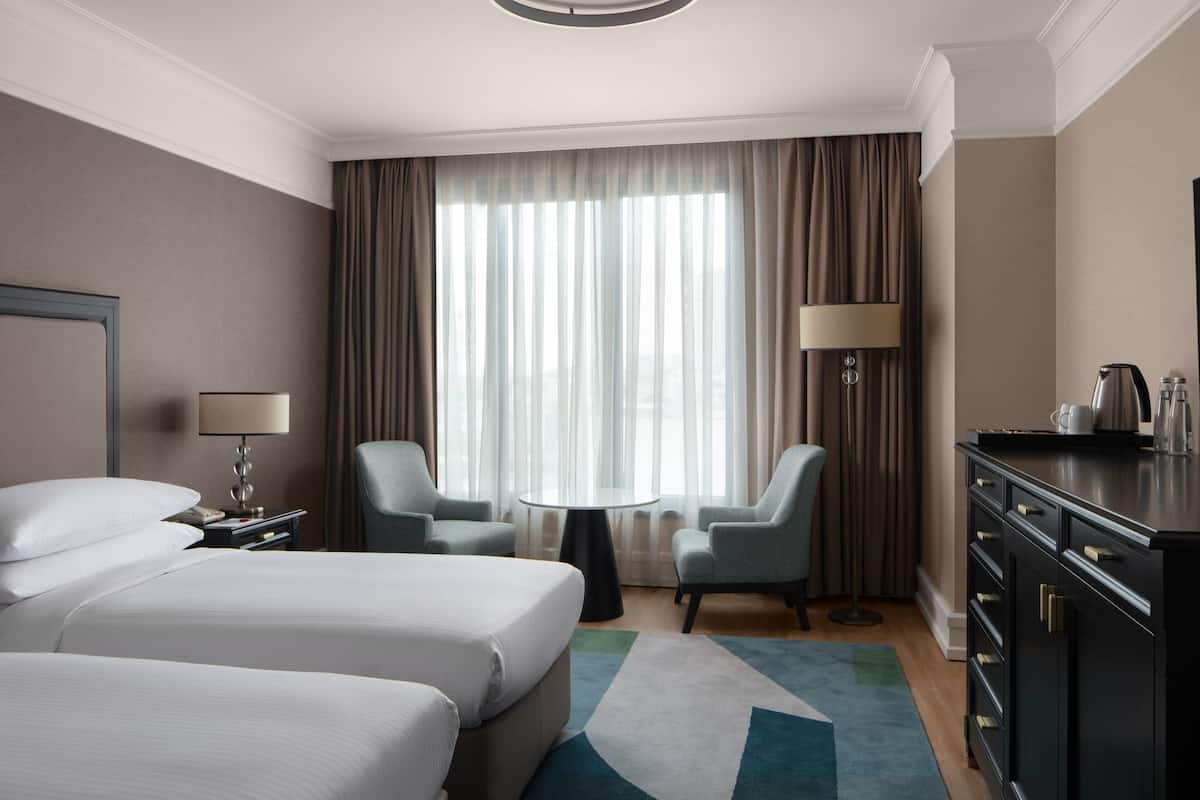 A hotel room with two beds, a desk, a chair, a lamp, a coffee maker, and a view of the city through large windows.