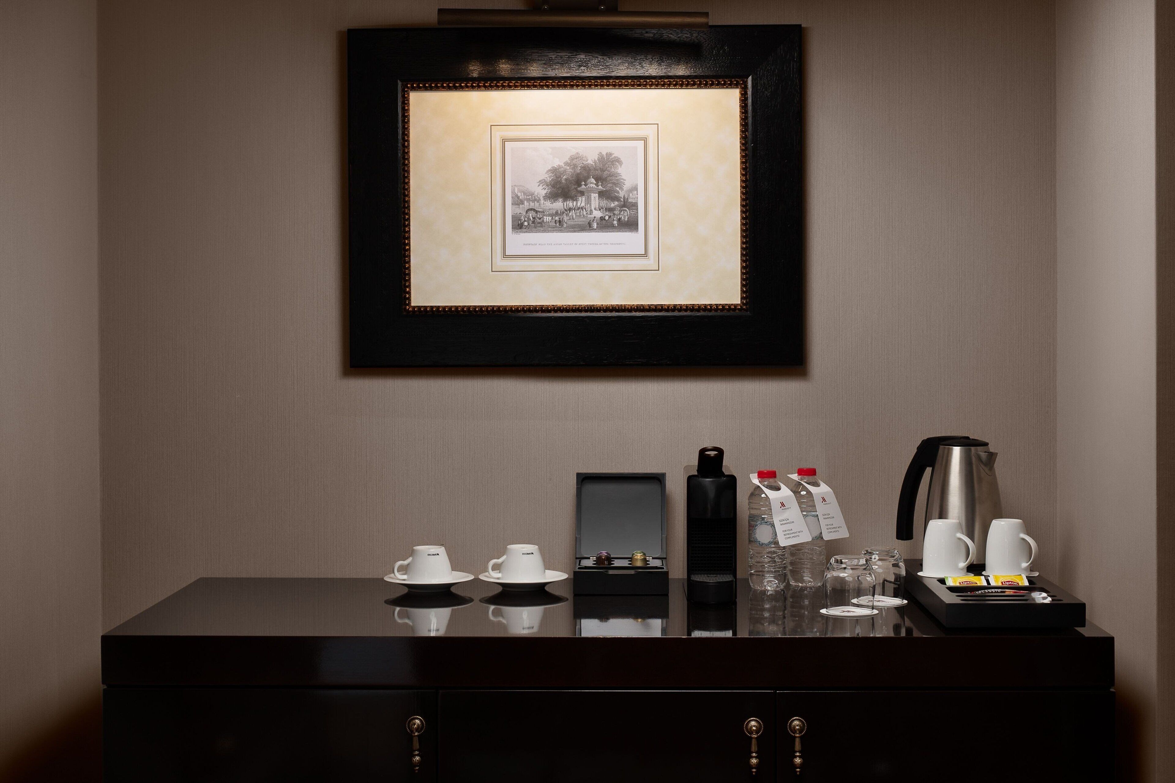 A hotel room with a framed picture on the wall, a dark wooden dresser with a coffee maker, cups, and a bottle of water.