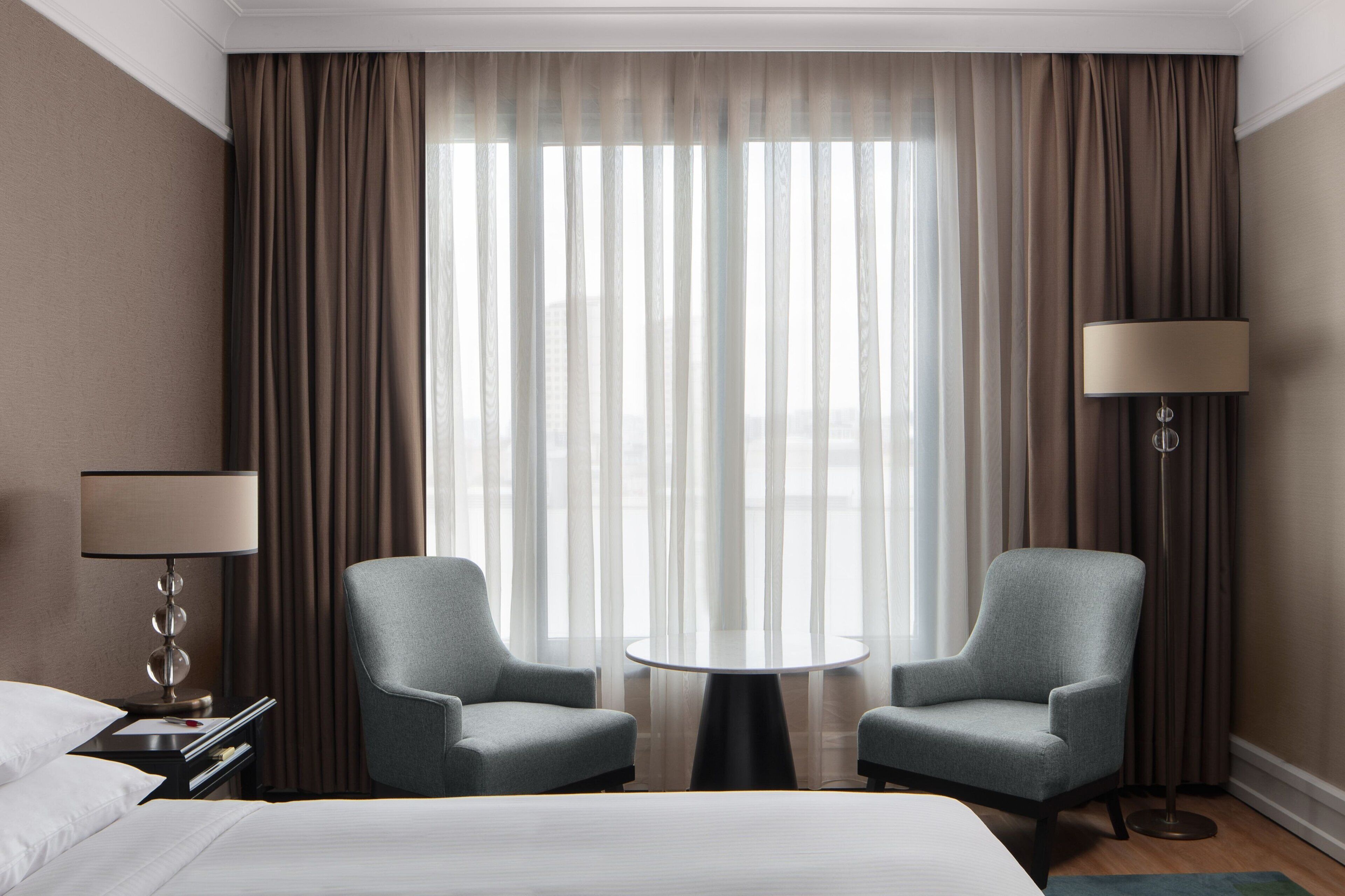 A hotel room with a bed, two armchairs, a small round table, a floor lamp, and a large window with sheer curtains.
