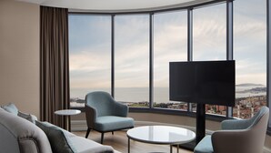 Una habitación de hotel moderna con un muro de vidrio curvo, un sofá, dos sillones, una pequeña mesa redonda y un televisor de pantalla plana.