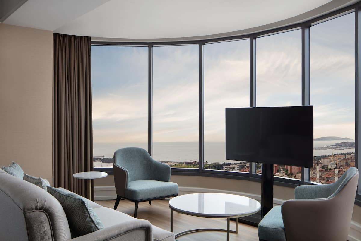 A modern hotel room with a curved glass wall, a sofa, two armchairs, a small round table, and a flat-screen TV.
