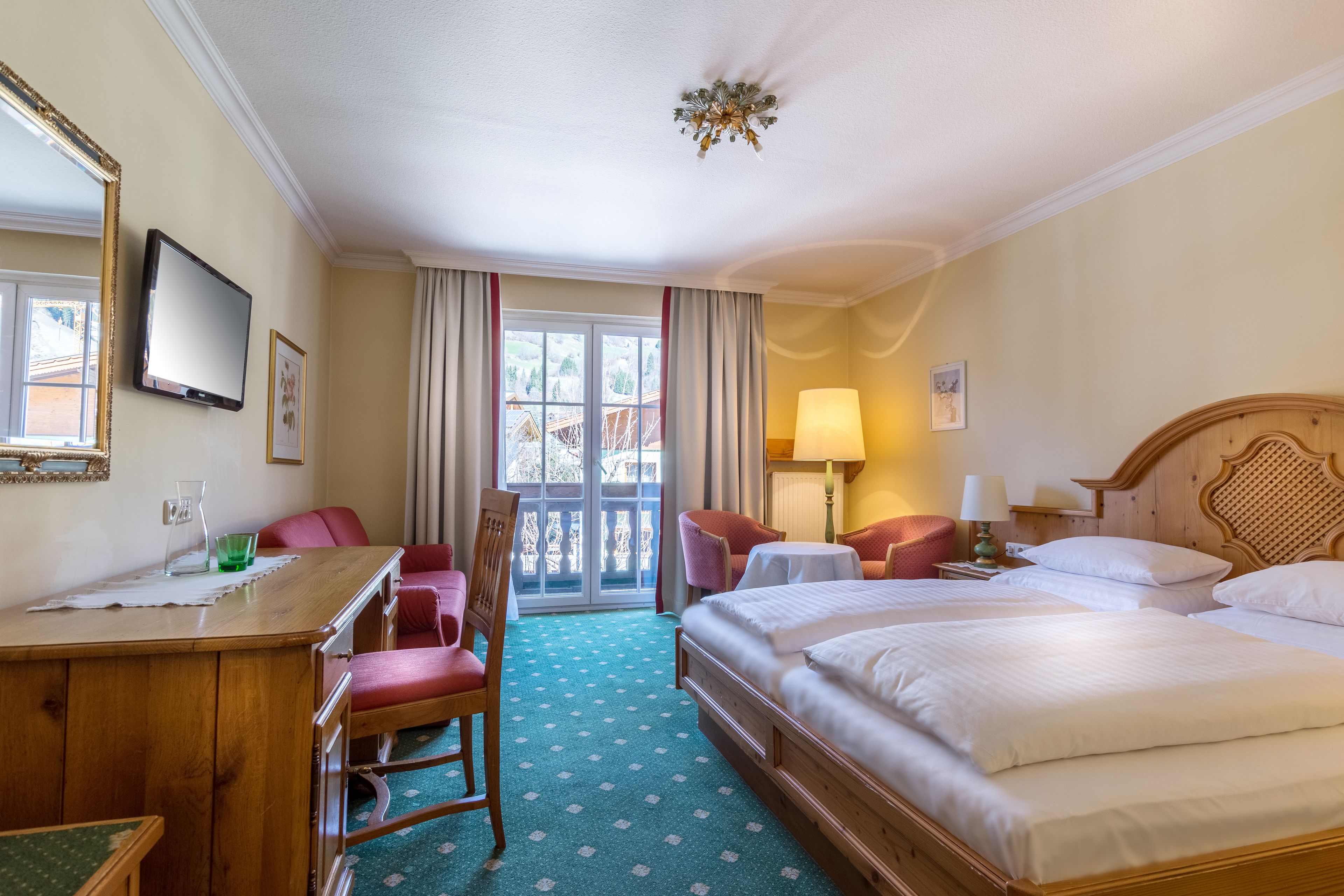Een hotelkamer met een houten bureau, twee eenpersoonsbedden, een stoel, een tafel en een balkon met gordijnen.