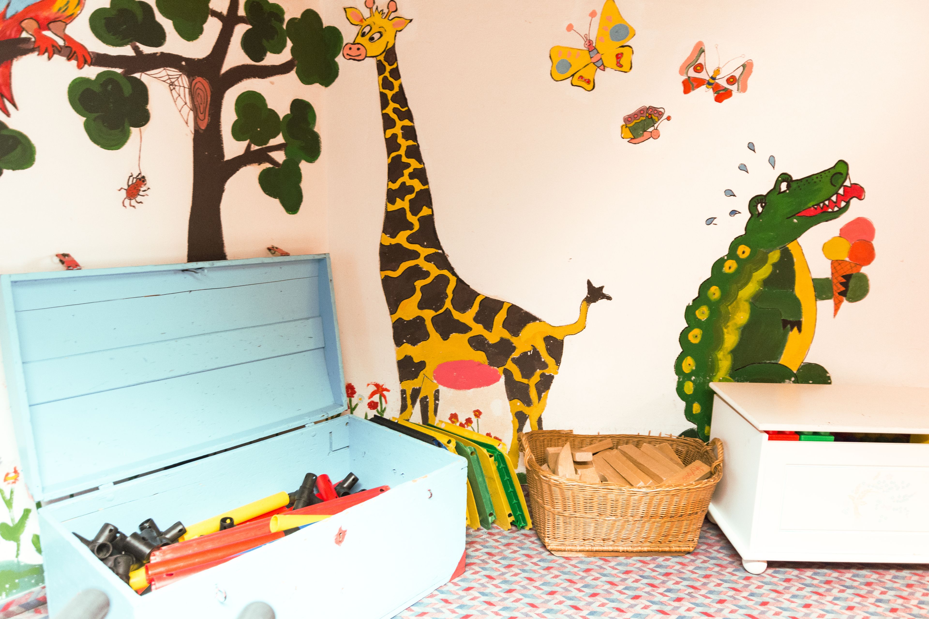 A child's room with a blue toy box, a white dresser, and a wall mural featuring a giraffe, a crocodile, and butterflies.