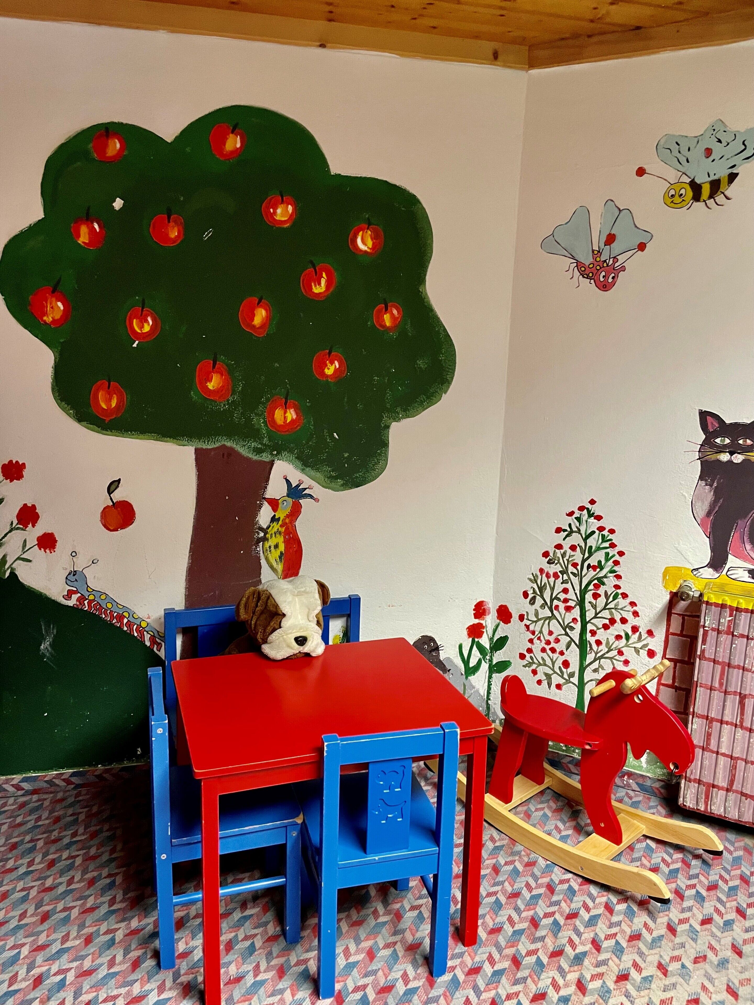 A child's room with a table, chairs, a rocking horse, and wall decorations including a tree, apples, and animals.