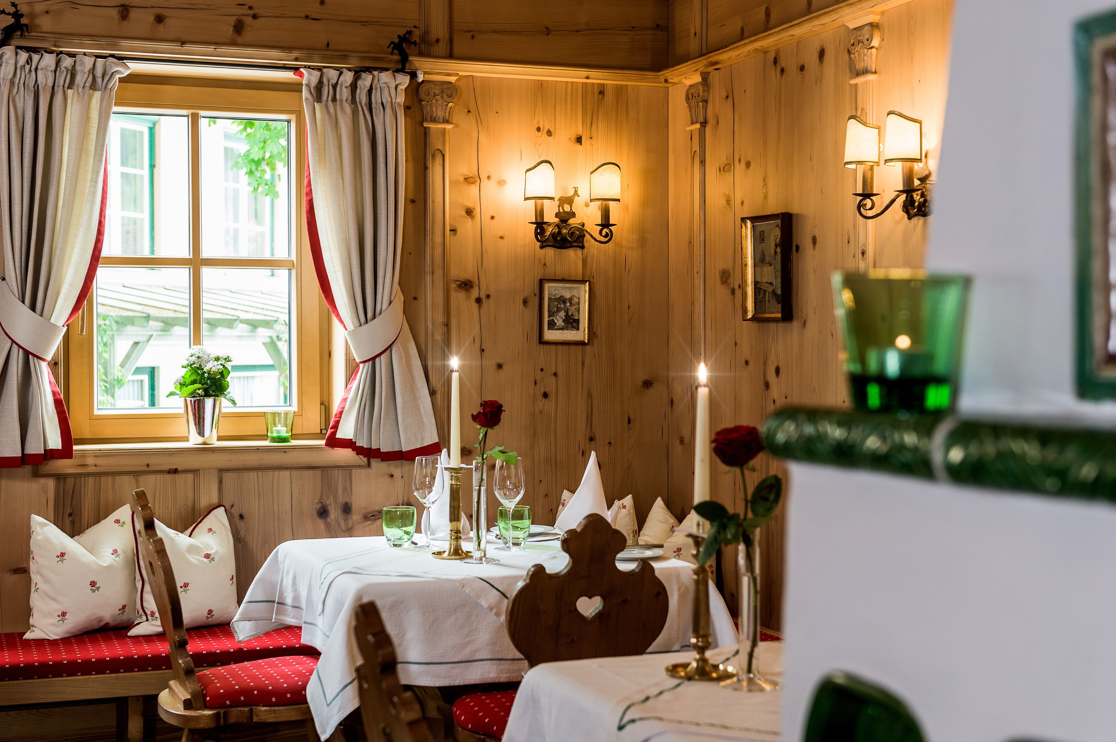A cozy dining area with wooden walls, a window with curtains, a table set with glasses and a vase of flowers, and wall-mounted lights.
