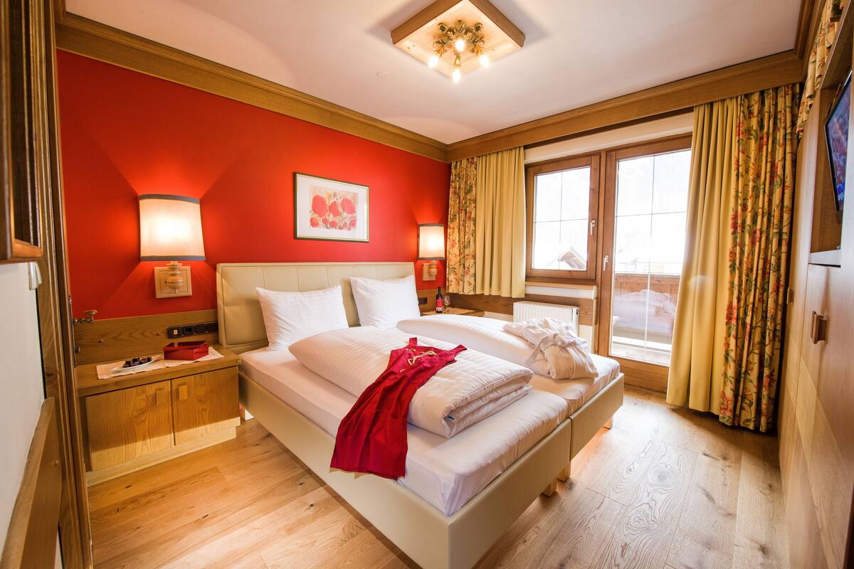 A hotel room with a large bed, two bedside tables, a red wall, and a window with curtains.
