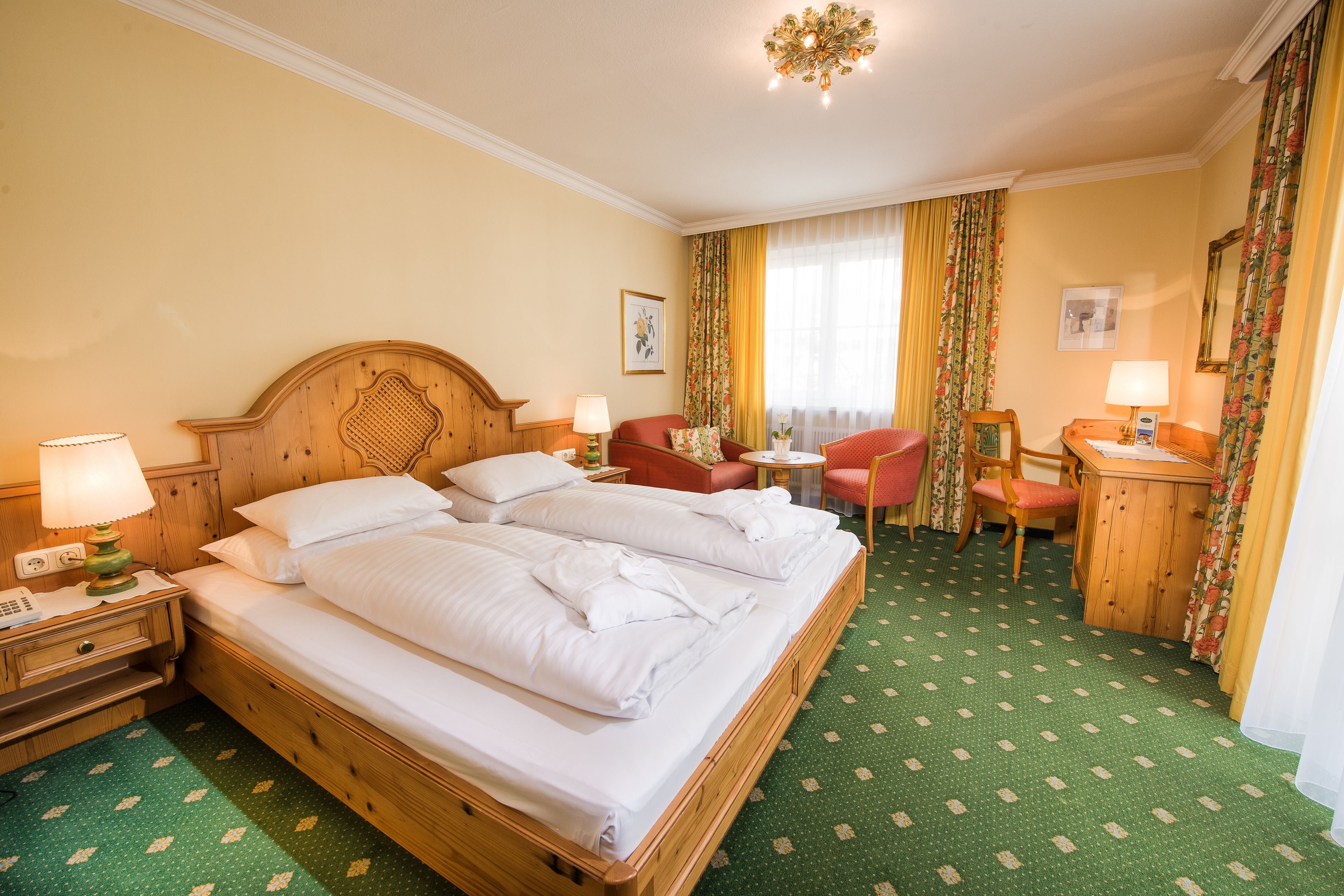 A hotel room with a large wooden bed, two bedside tables with lamps, a small desk with a chair, and a green patterned carpet.