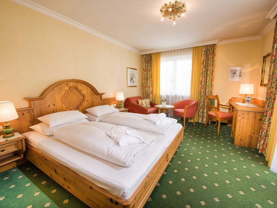 A hotel room with a large wooden bed, two bedside tables with lamps, a small desk with a chair, and a green patterned carpet.
