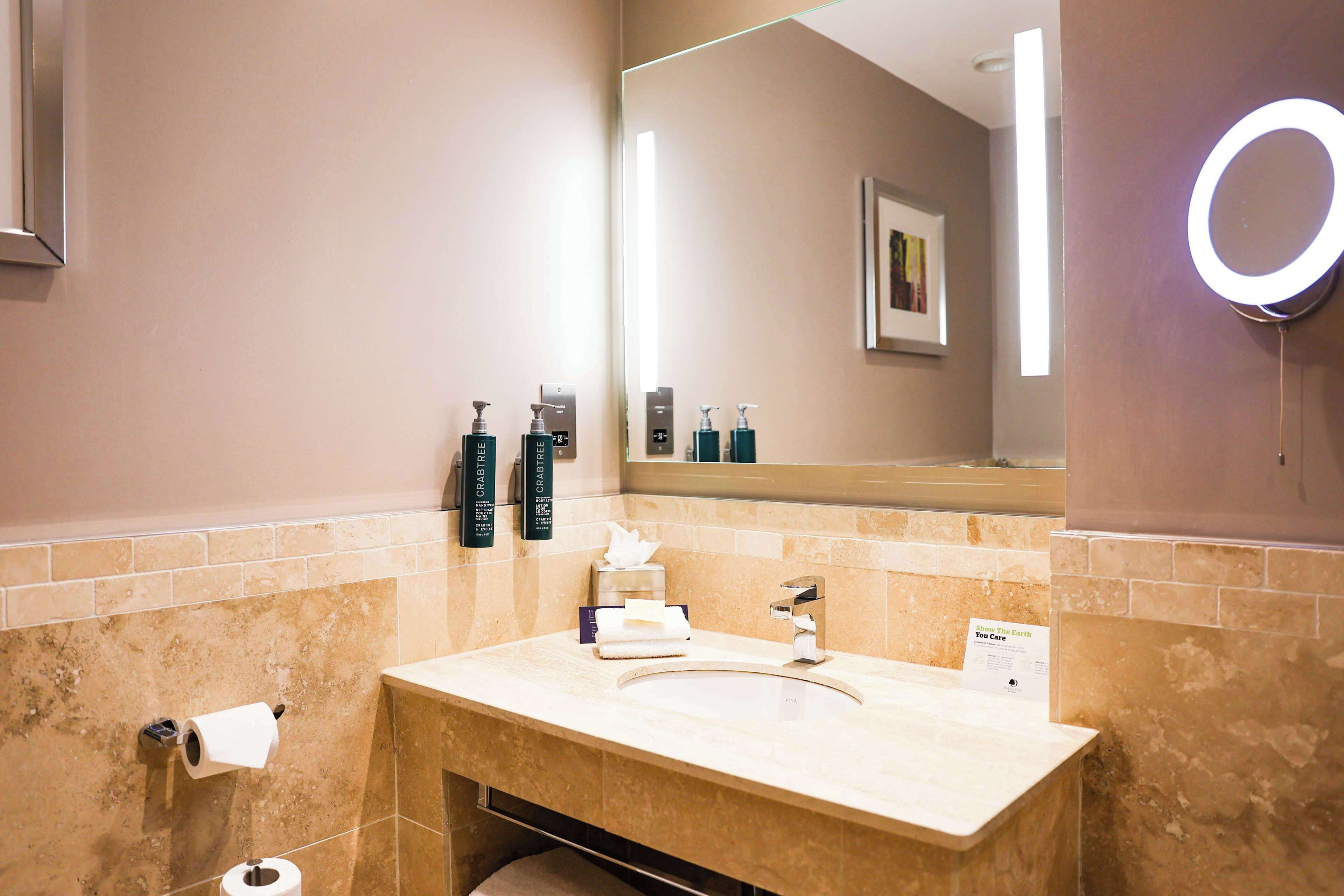 A bathroom with a marble sink, a mirror with a light, and toiletries on the wall.