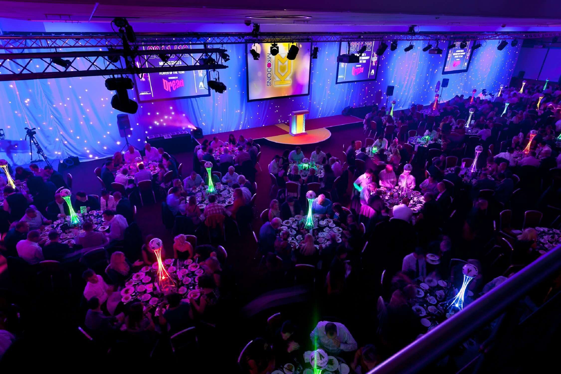 A crowded event space with tables and chairs arranged for an audience, a stage with a podium, and a backdrop with text and graphics.