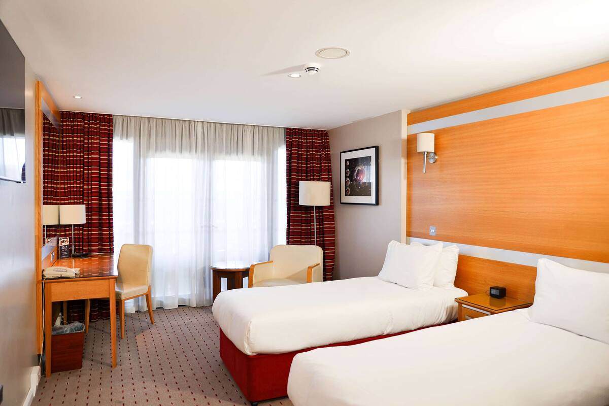 Une chambre d’hôtel avec deux lits, un bureau, une chaise, une petite table et une lampe.
