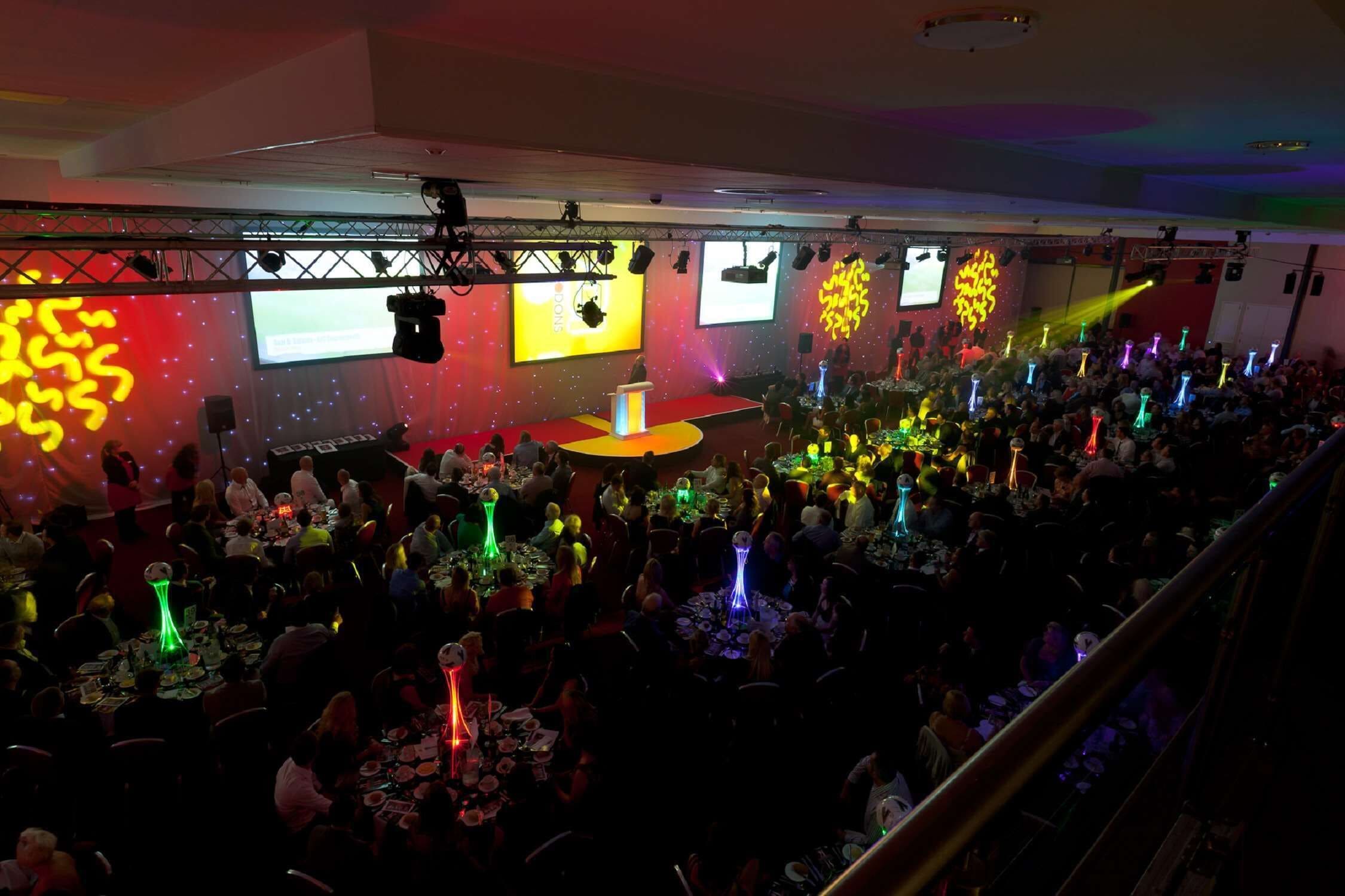 Evento concurrido en interior, con escenario, mesas y asistentes.