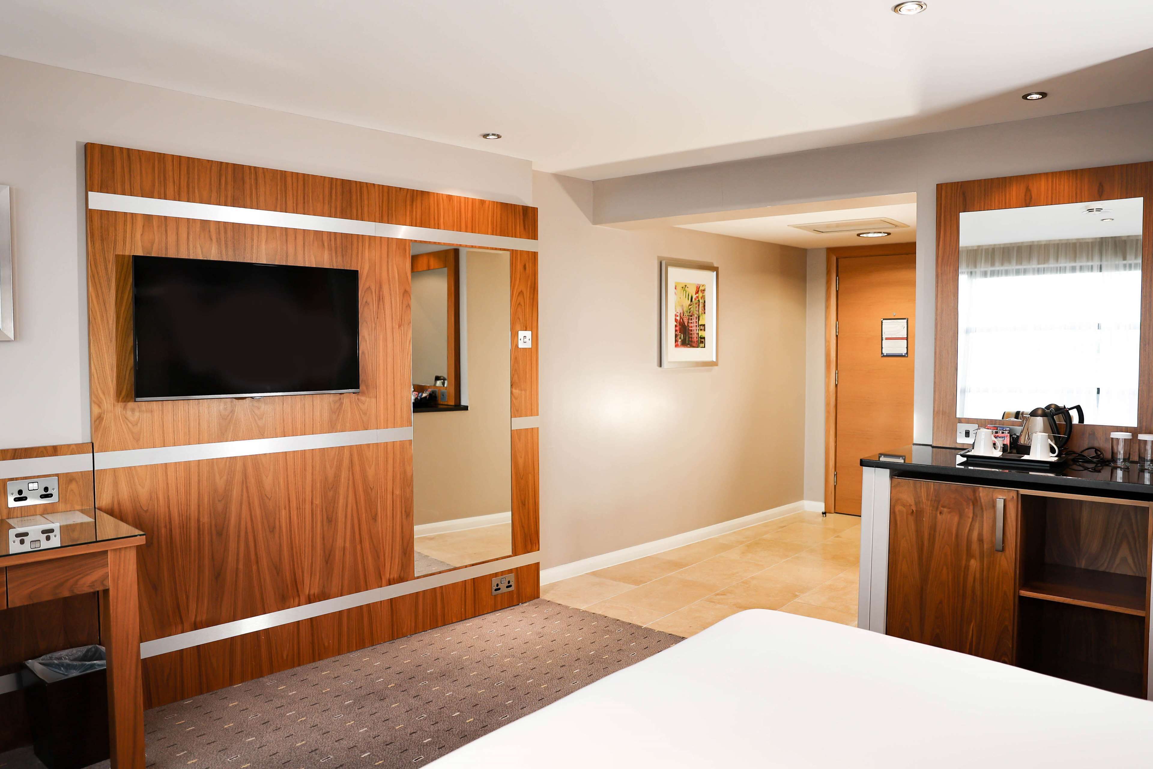 A modern hotel room with a large flat-screen TV mounted on a wooden panel, a small desk with a trash bin, a bed with a white mattress, and a wooden wardrobe with a mirror.