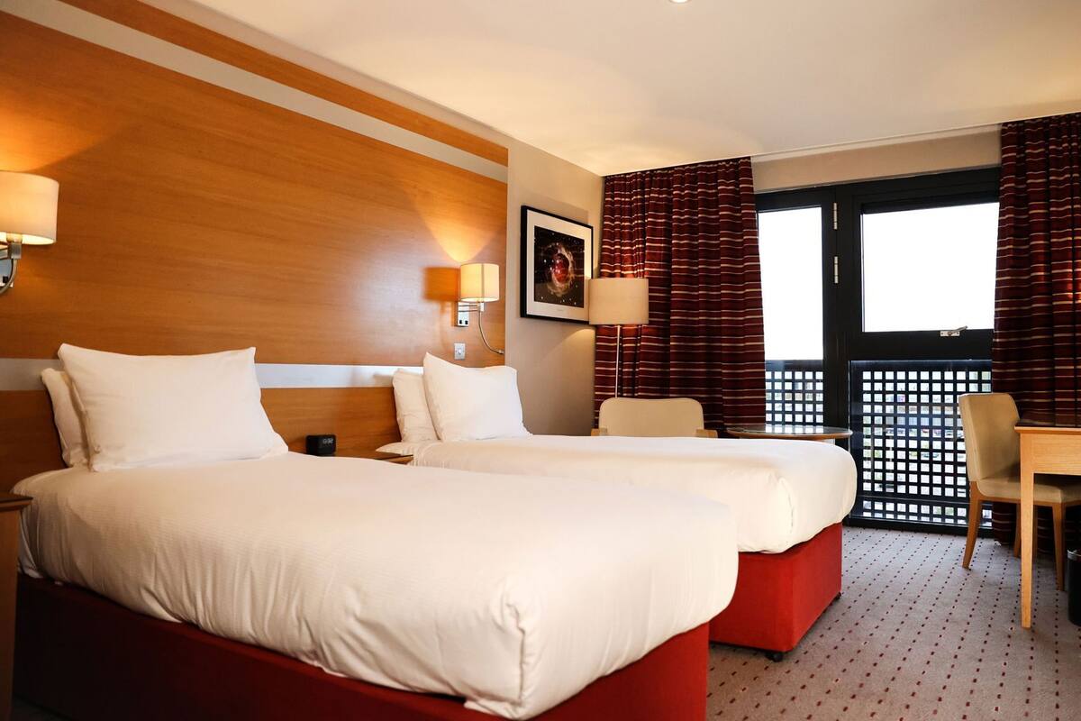 A hotel room with two beds, a desk, a chair, and a window with curtains.