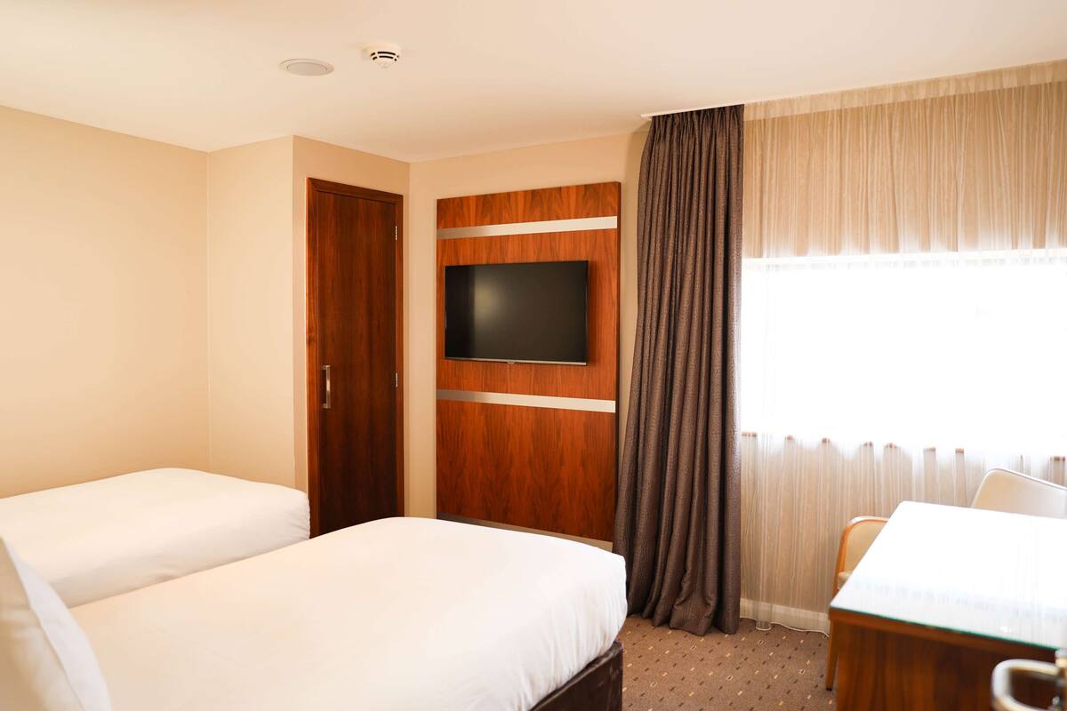 A hotel room with two beds, a TV mounted on a wooden panel, a dining table, and a window with curtains.