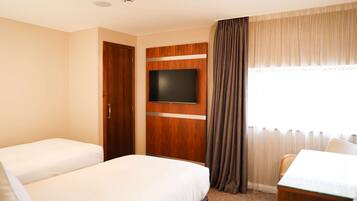 A hotel room with two beds, a TV mounted on a wooden panel, a dining table, and a window with curtains.