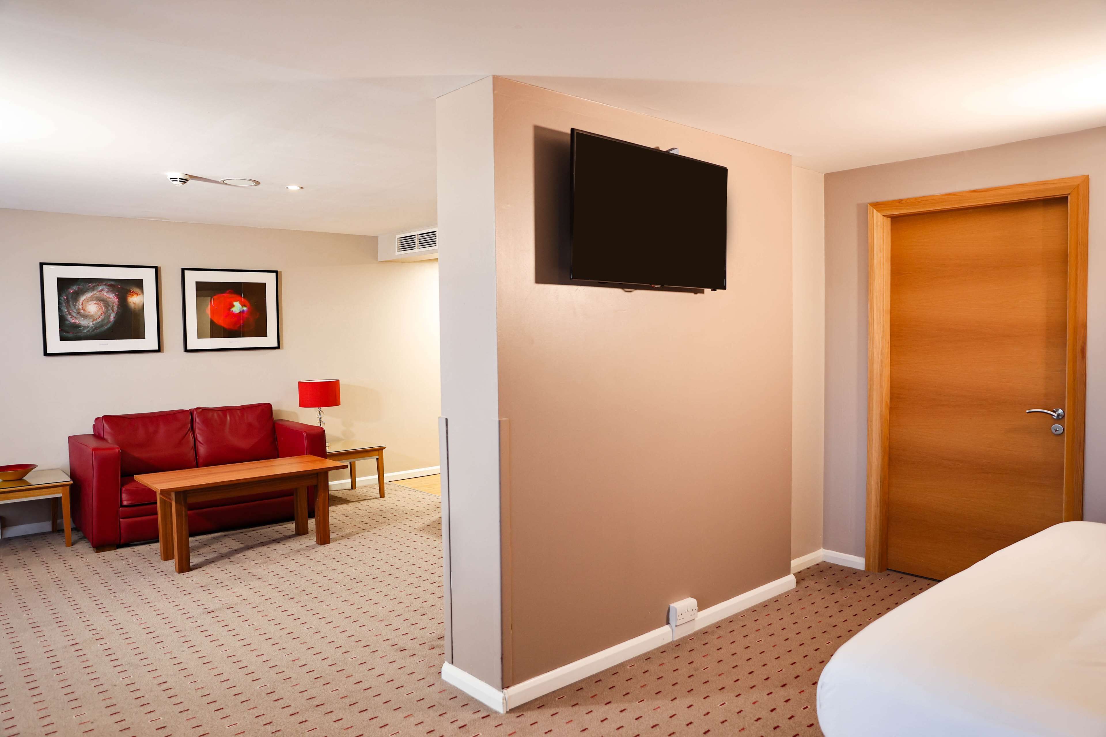 Une chambre d’hôtel avec un lit, un canapé en cuir rouge, une table basse en bois, un téléviseur fixé au mur et deux tableaux encadrés accrochés au mur.