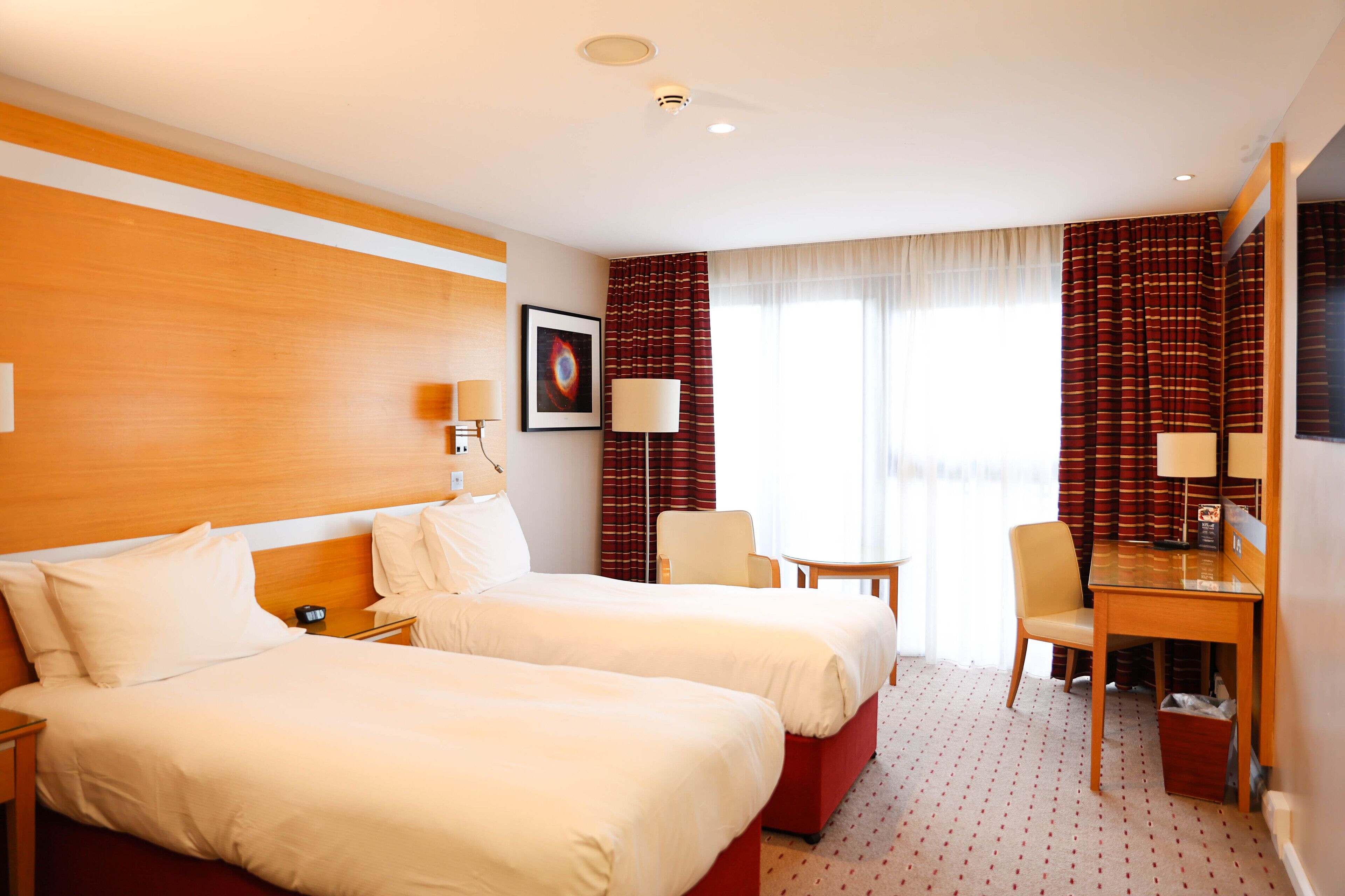 A hotel room with two beds, a desk, a chair, a lamp, and a framed picture on the wall.