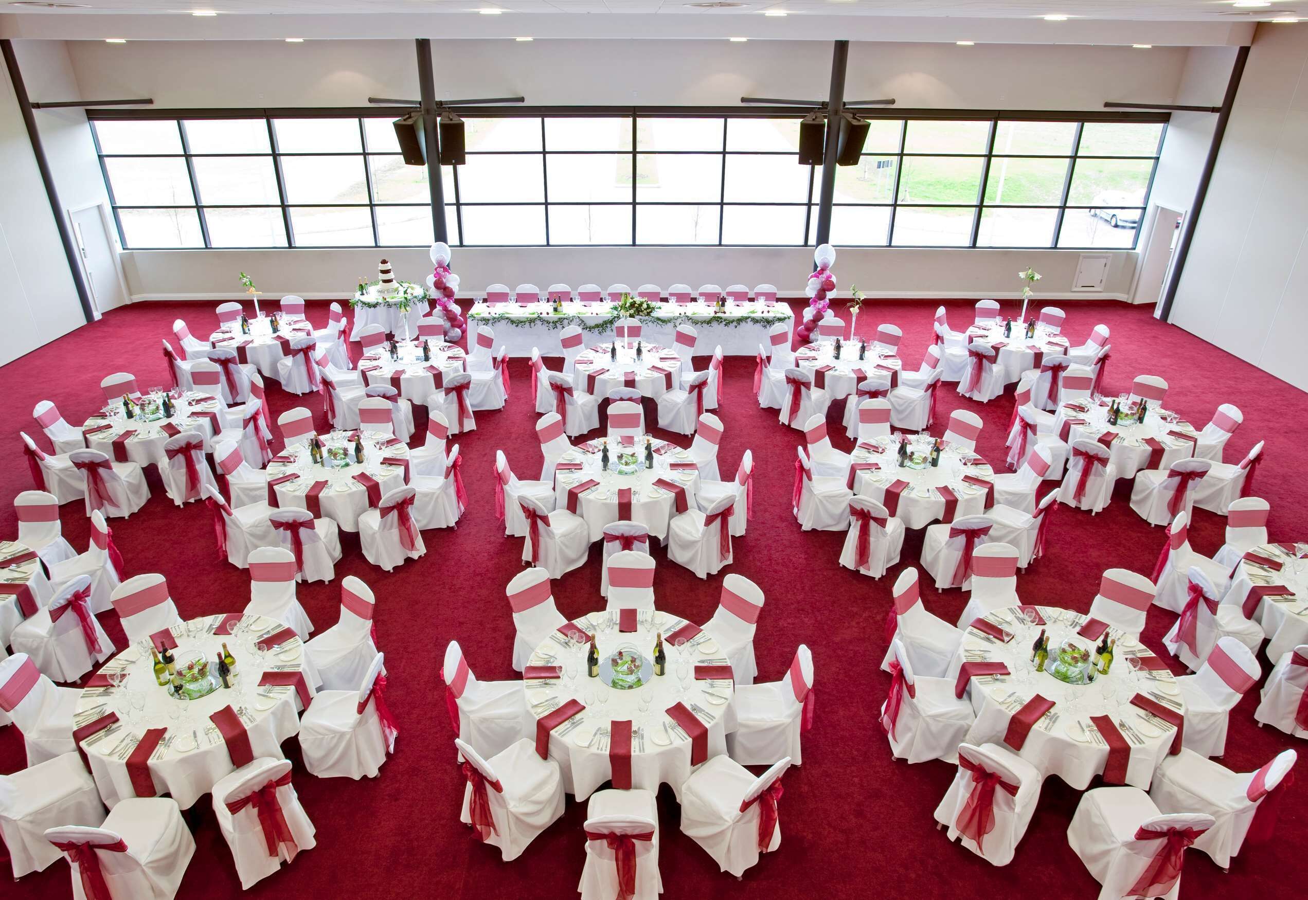 A large hall with multiple round tables, each with white tablecloths and pink sashes, arranged in a circular pattern.