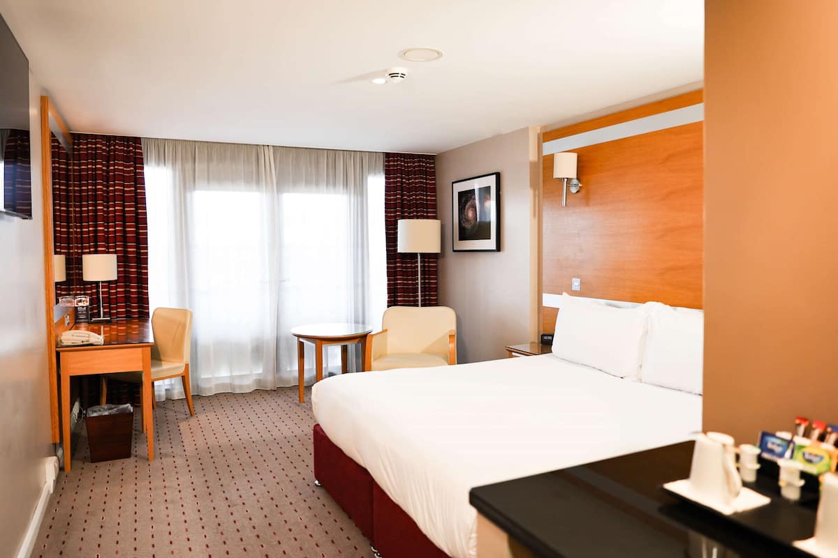 Une chambre d’hôtel avec un lit, un bureau et une chaise, une petite table, une lampe et un tableau au mur.