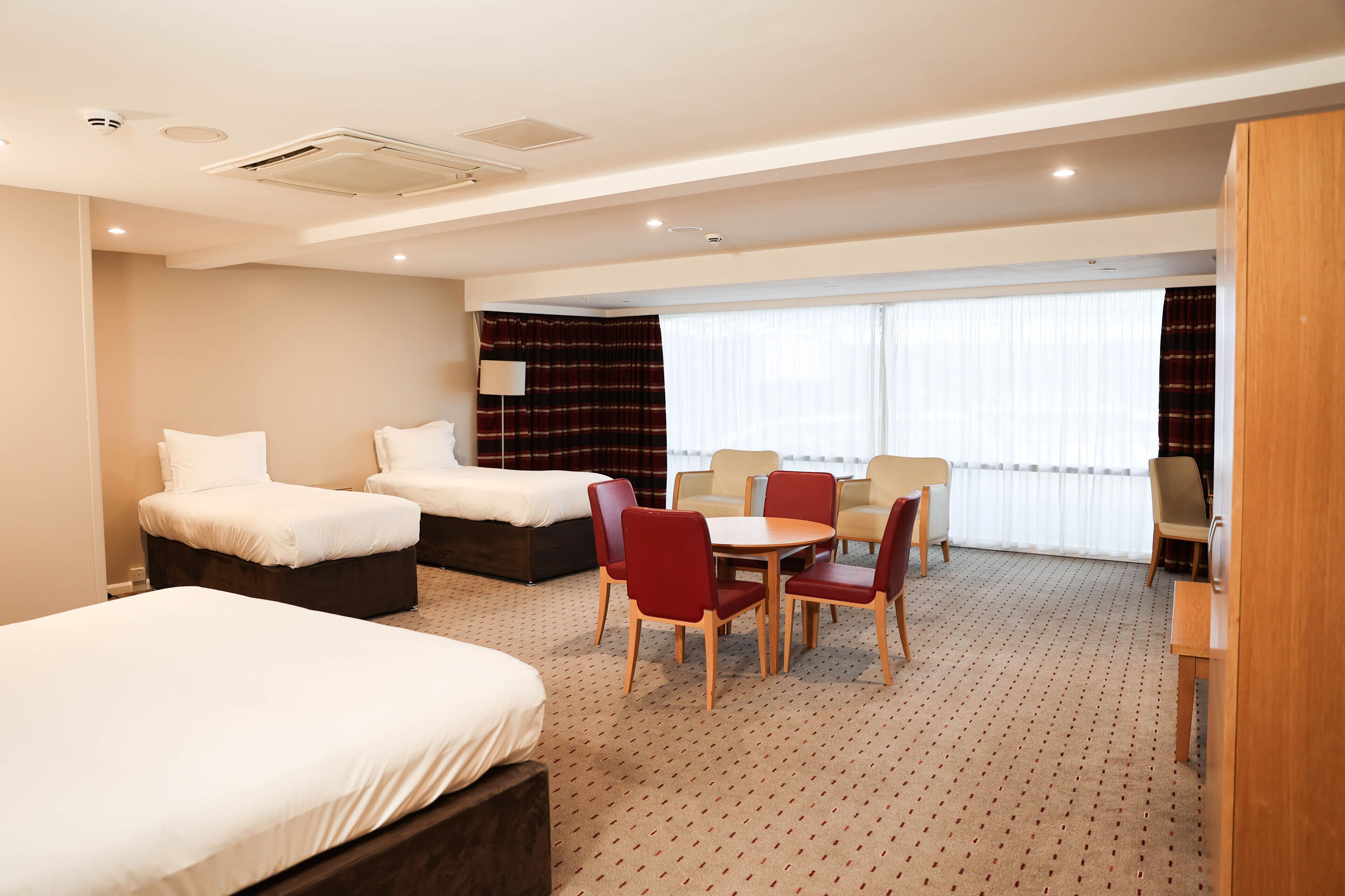 Ein Hotelzimmer mit zwei Betten, einem Esstisch mit Stühlen, einer kleinen Küchenzeile und einem großen Fenster mit Vorhängen.
