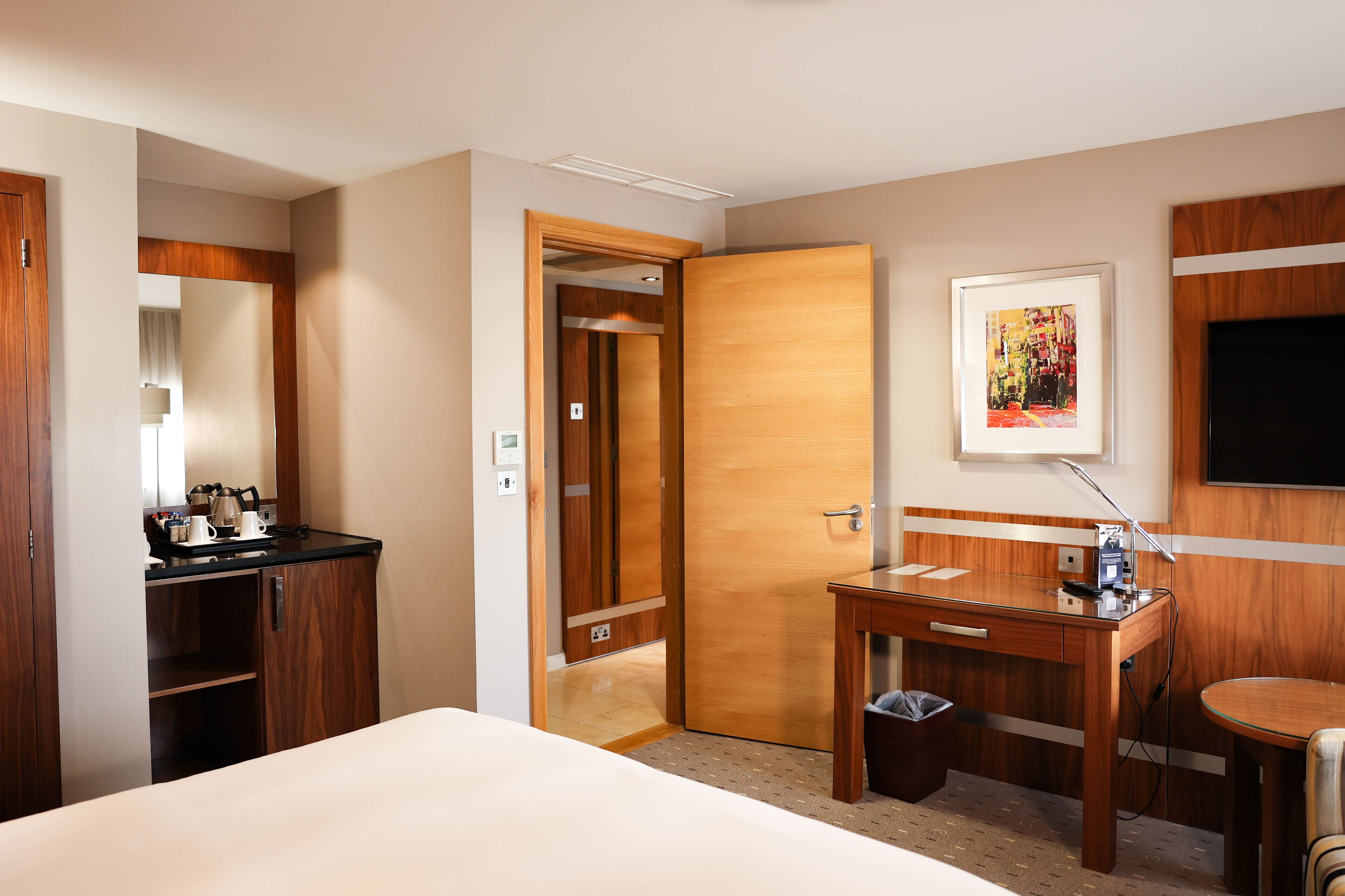 A hotel room with a bed, a desk with a chair, a small table, a TV, and a door leading to a bathroom.
