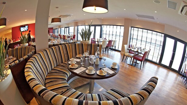 A modern dining area with a curved sofa and a round table set for a meal, large windows offering a view, and a television mounted on the wall. - DoubleTree by Hilton Milton Keynes (Milton Keynes)