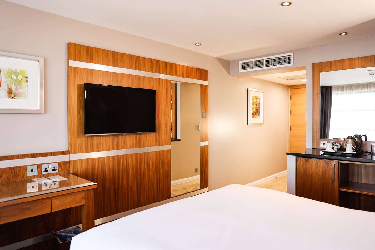 A modern hotel room with a wooden panel wall, a flat-screen TV, a desk with a mirror, and a kitchenette area.