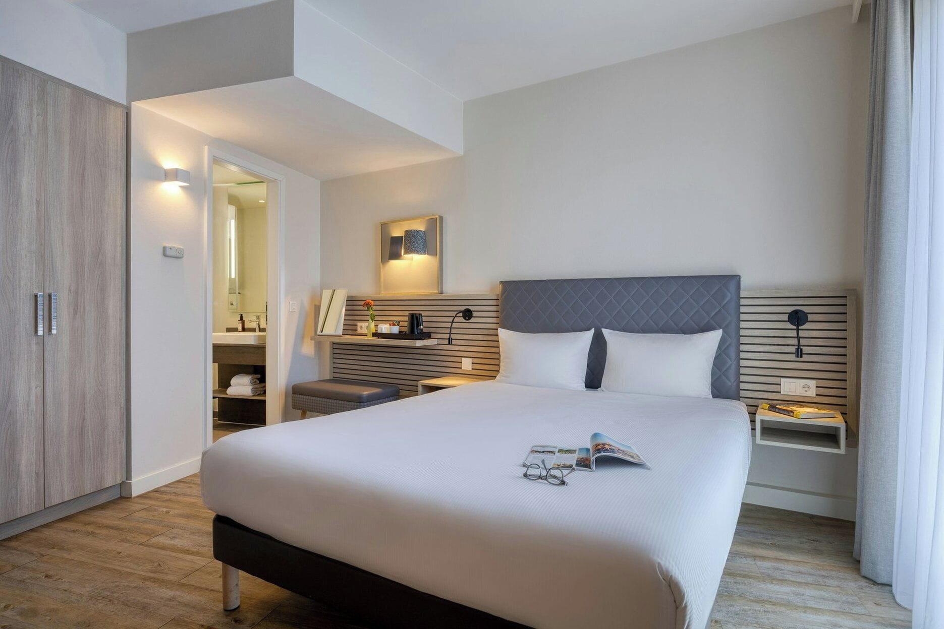 A modern hotel room with a large bed, bedside tables, a wardrobe, and a bathroom visible through an open door.