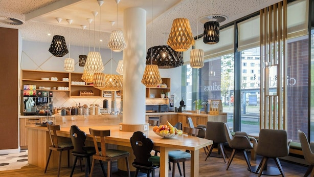 A modern cafe with a wooden table, chairs, a coffee machine, and hanging pendant lights. - Citadines Arnulfpark Munich (Munich)