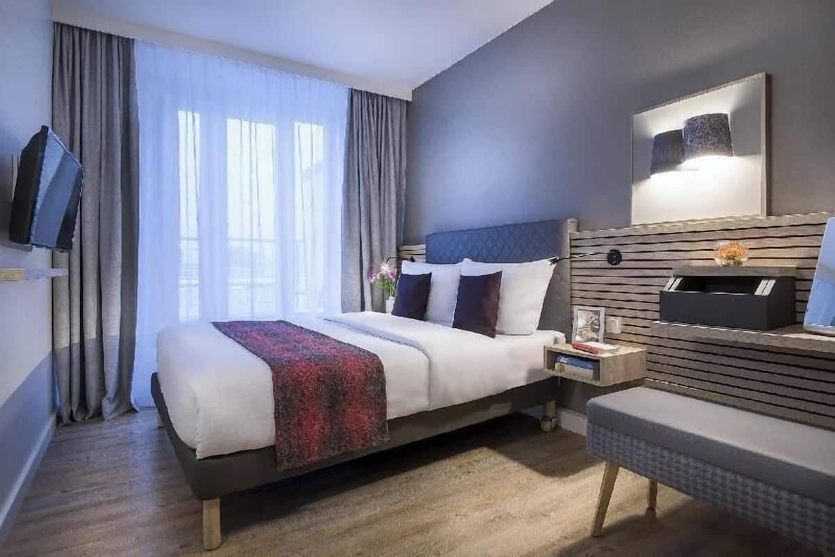 A hotel room with a bed, a desk, a chair, a TV, and a large window with curtains.