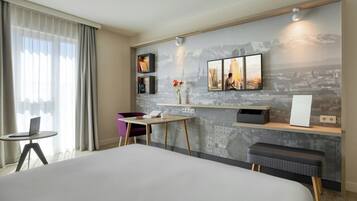 A hotel room with a large bed, a desk with a laptop, a small table with a vase, and a wall-mounted TV.